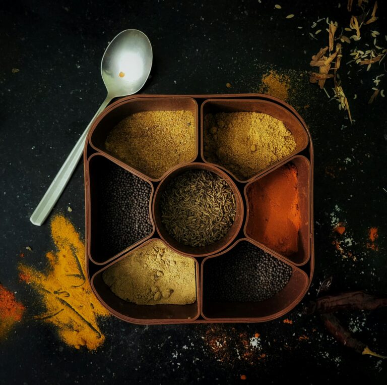 A top view of a spice box filled with various Indian spices, surrounded by scattered colorful powders.